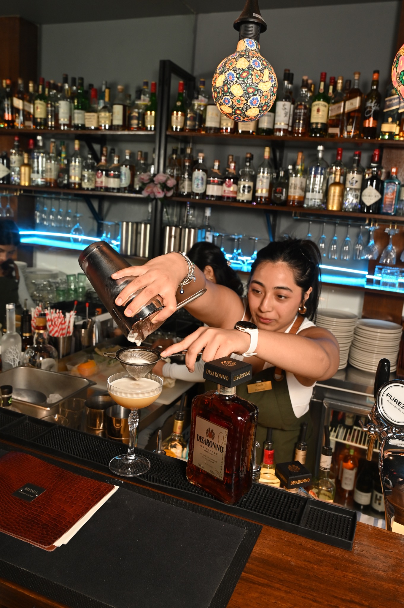 A bartender straining a cocktail into a coupe beneath a colourful mosaic lamp at Bombay on Bay