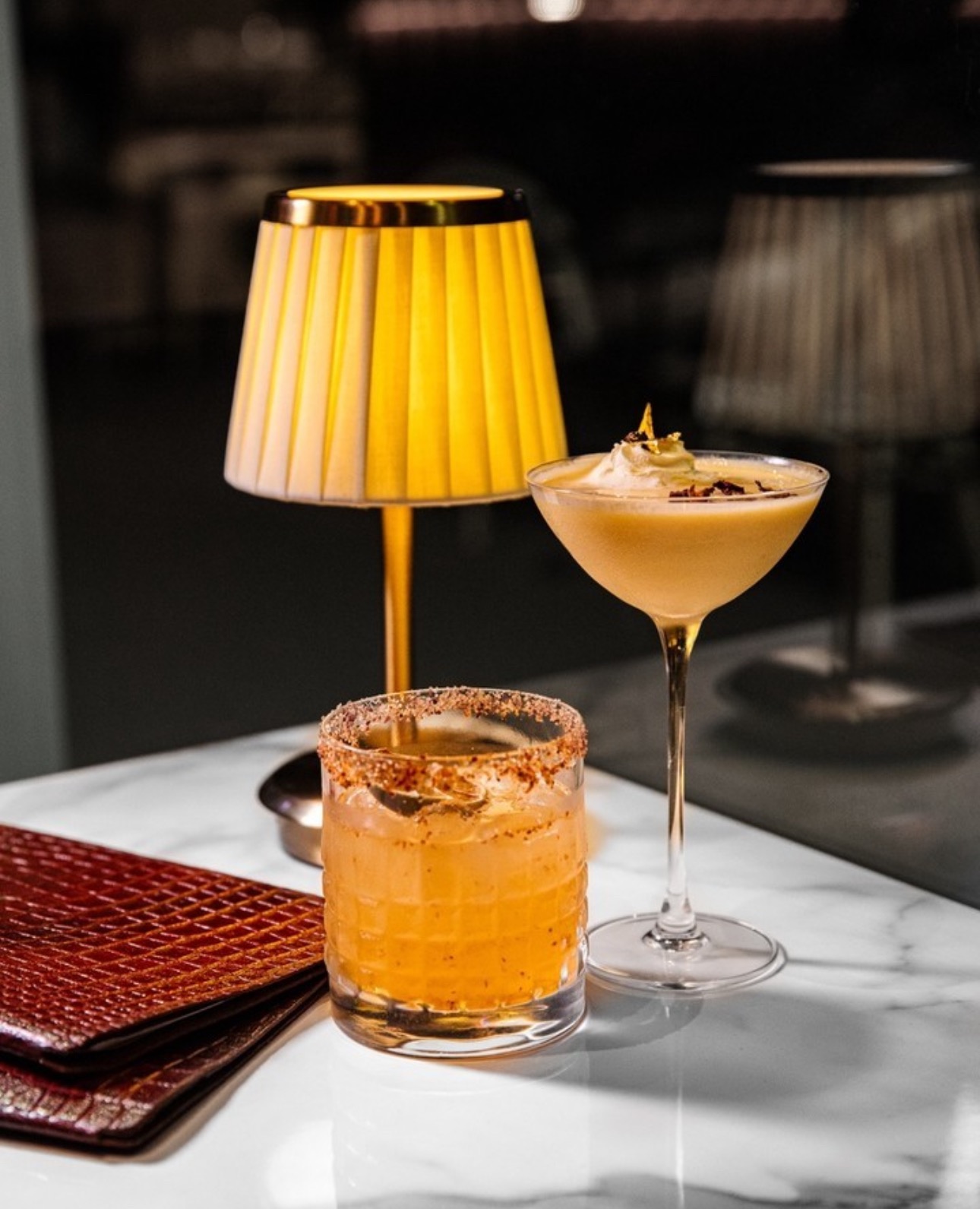 Two house cocktails on a marble table beside a glowing golden lamp